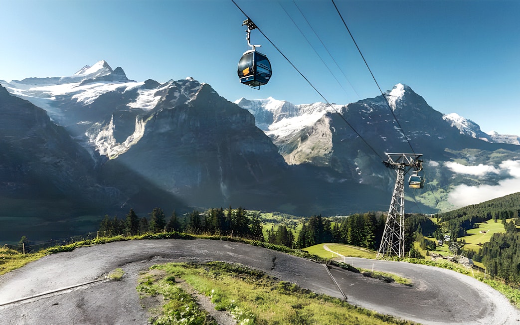 Cable cars over paths with snow-covered peaks and green valleys at Grindelwald First, Swiss Alps.