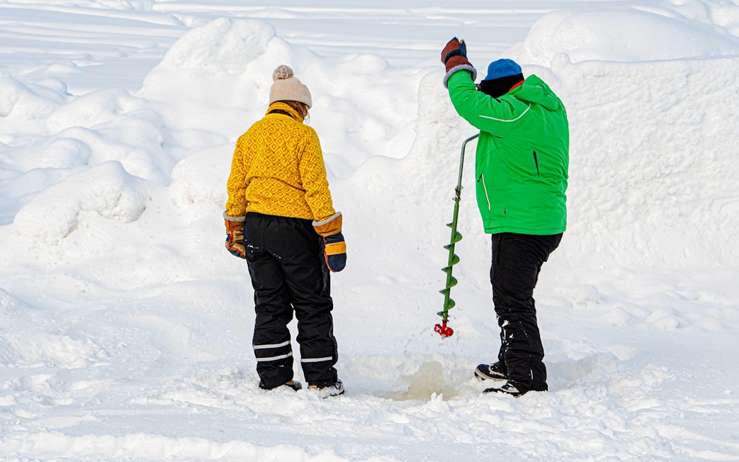Two people drilling a hole in the ice for fishing in a snowy landscape.