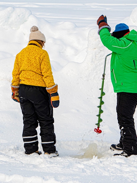 Two people drilling a hole in the ice for fishing in a snowy landscape.