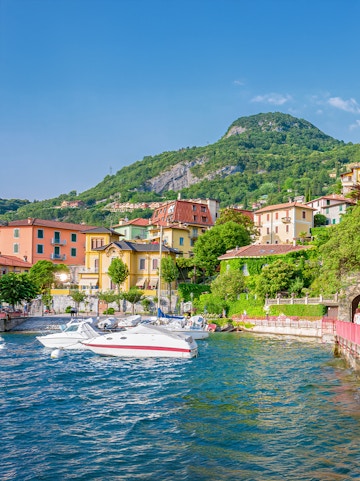 Boats docked by colorful buildings on Lake Como, Italy, with lush hills in the background.