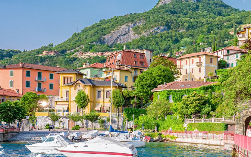 Boats docked by colorful buildings on Lake Como, Italy, with lush hills in the background.