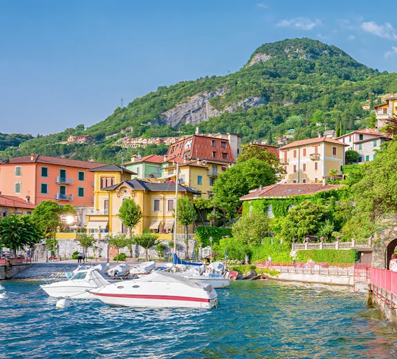 Boats docked by colorful buildings on Lake Como, Italy, with lush hills in the background.