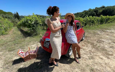 Women enjoying wine by a red Fiat 500 in a Tuscan vineyard during a scenic drive.
