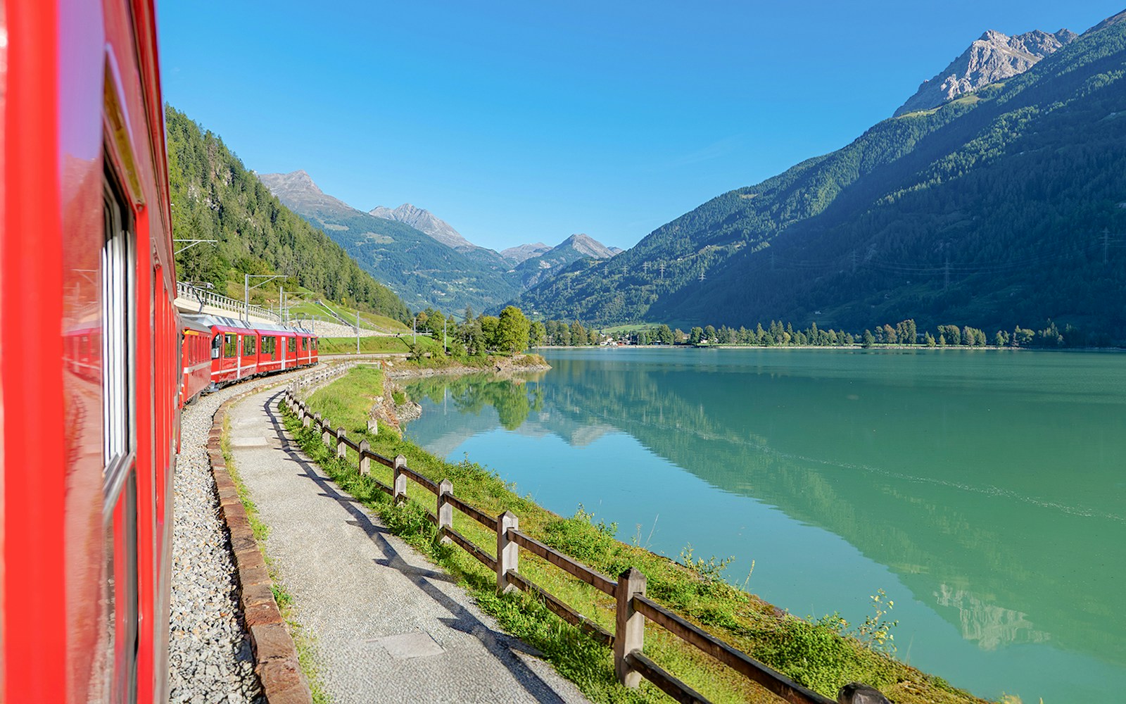 Red train of Bernina passing through scenic Poschiavo, Switzerland, with snow-capped mountains in the background.