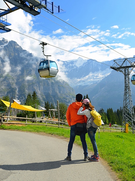 Couple embracing near cable cars at Grindelwald First, Switzerland, with mountain views.