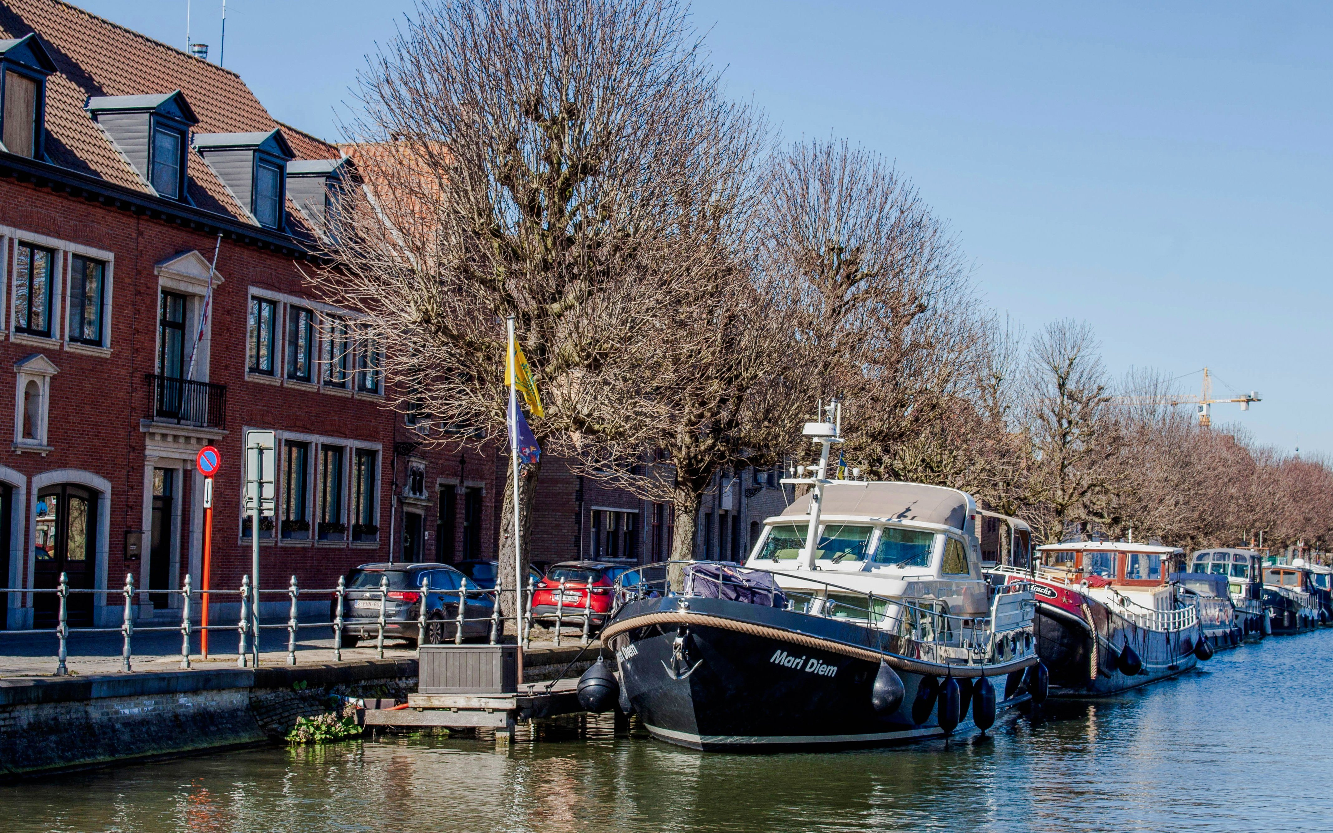 Boats moored along Coupure canal in Bruges with brick buildings and bare trees.