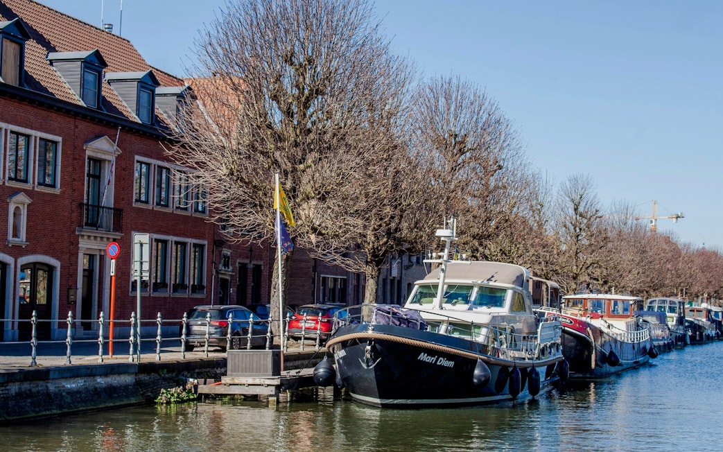 Boats moored along Coupure canal in Bruges with brick buildings and bare trees.