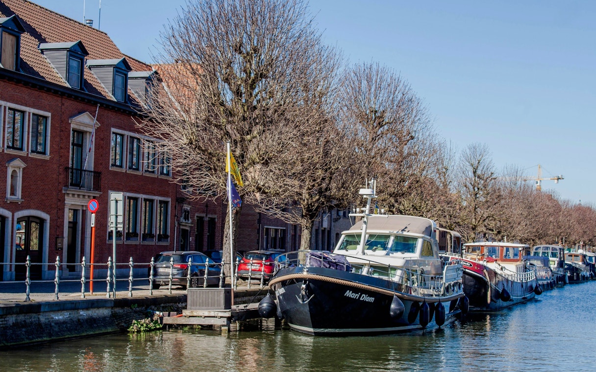 Boats moored along Coupure canal in Bruges with brick buildings and bare trees.