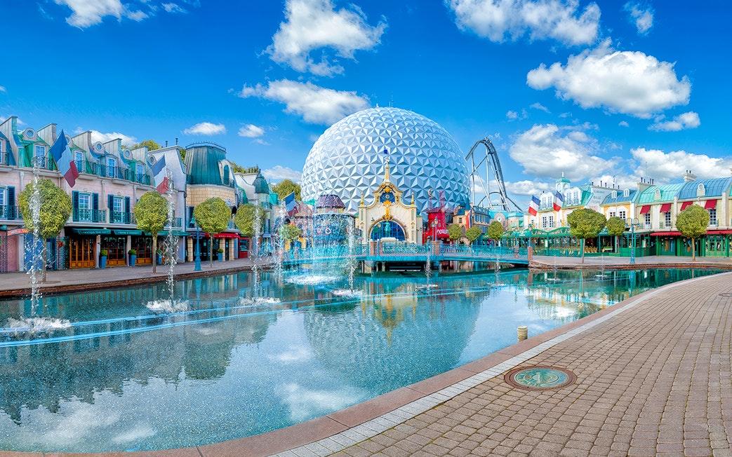 Theme park with geodesic dome, fountains, and colorful buildings reflecting in water.