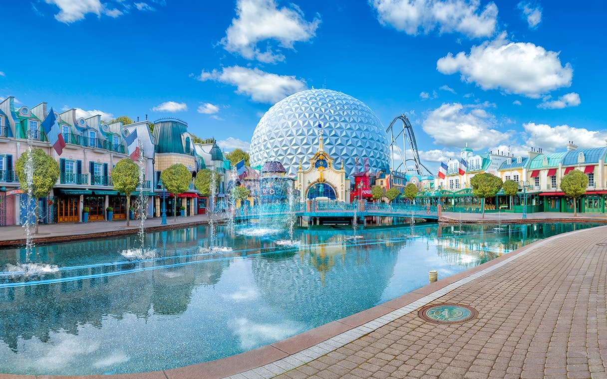 Theme park with geodesic dome, fountains, and colorful buildings reflecting in water.