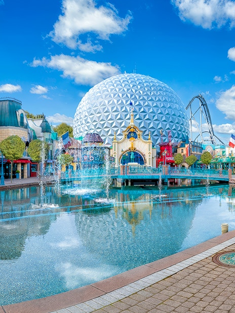 Theme park with geodesic dome, fountains, and colorful buildings reflecting in water.