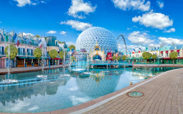Theme park with geodesic dome, fountains, and colorful buildings reflecting in water.