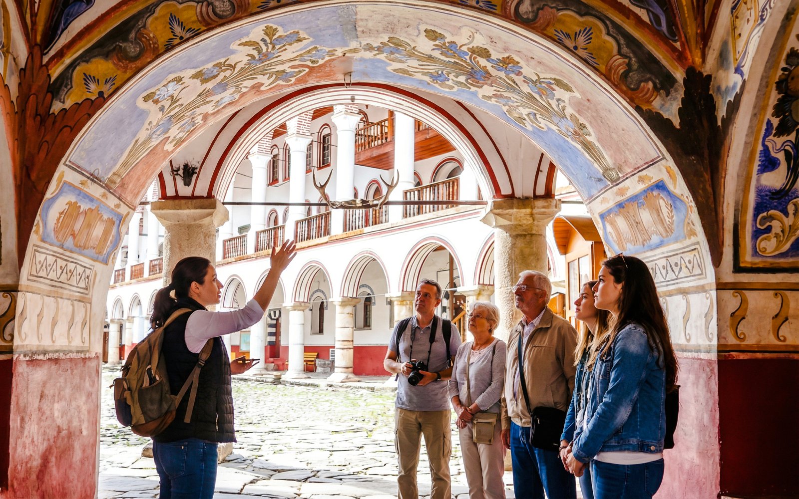 Tour guide explaining to visitors under ornate arch at Rila Monastery, Bulgaria.