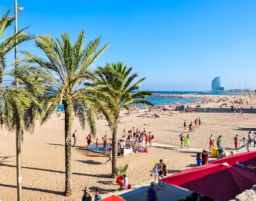 Tourists at Barceloneta Beach, Barcelona, with palm trees and sea view.