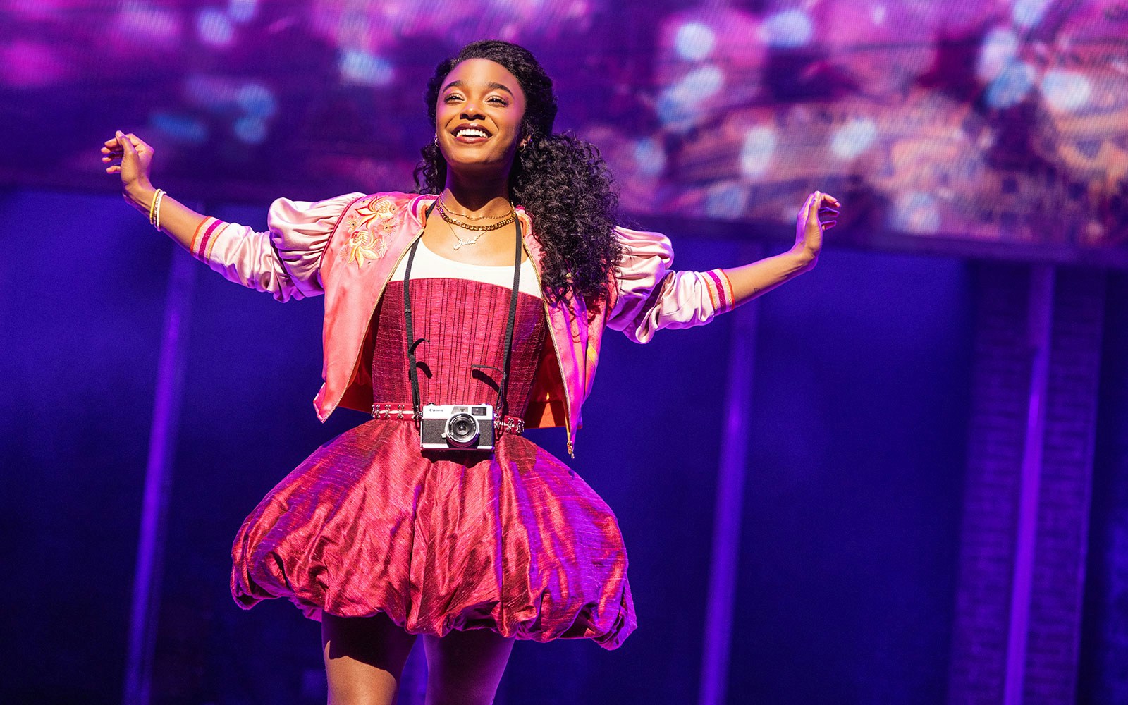 Performer on stage in & Juliet musical, wearing a pink dress and holding a camera.