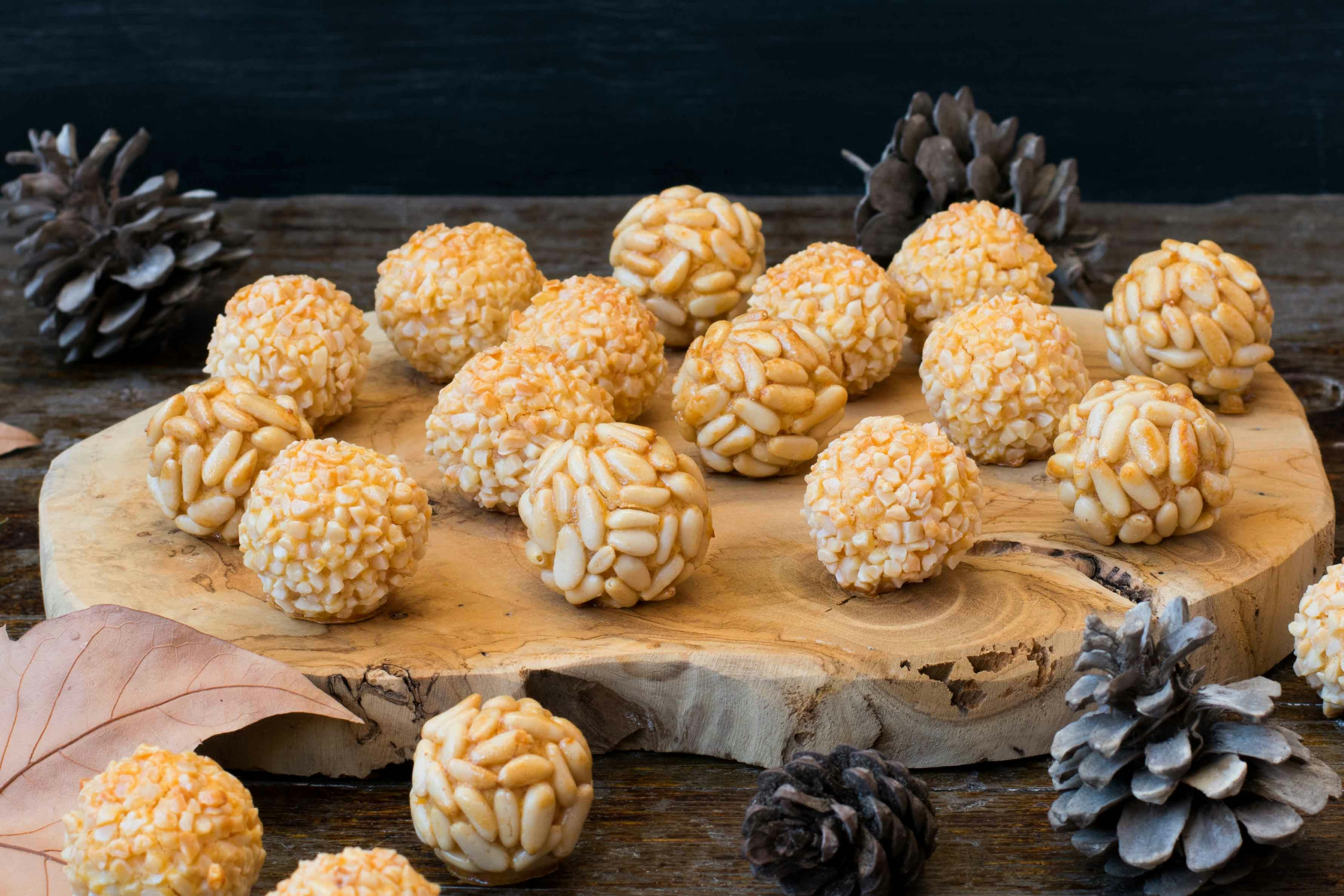 Panellets with pine nuts on a wooden board surrounded by pine cones.