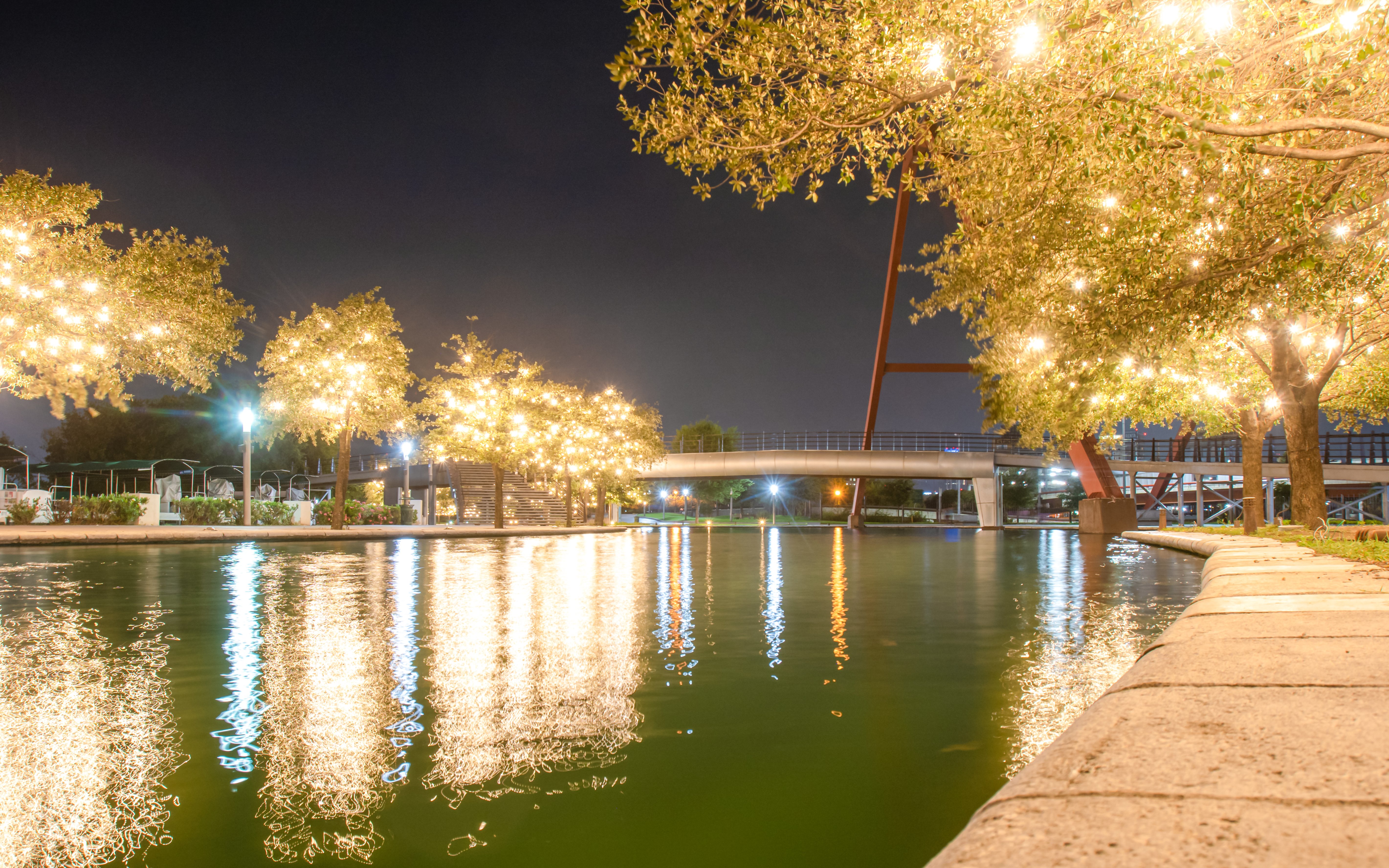 Lights reflecting on canal water at night with illuminated trees and a bridge.