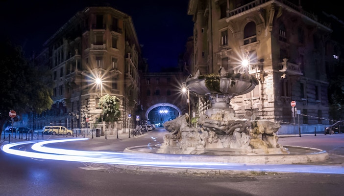 Fountain in Coppedè district, Rome, illuminated at night with light trails from passing cars.