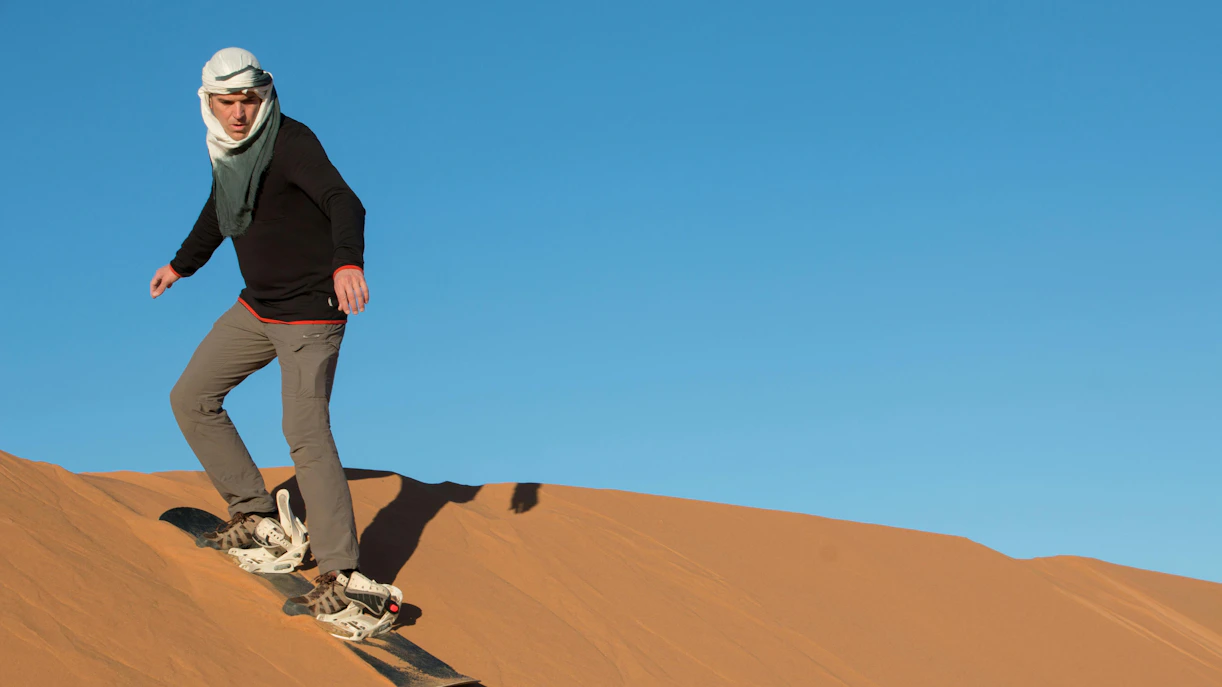 Tourist sandboarding down a desert dune under a clear blue sky.