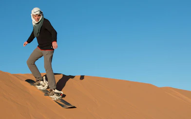 Tourist sandboarding down a desert dune under a clear blue sky.