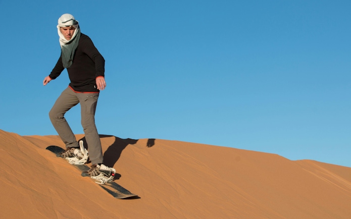 Tourist sandboarding down a desert dune under a clear blue sky.