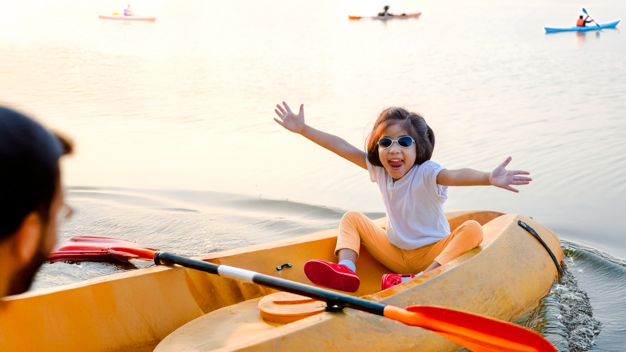 Toddler kayaking on a calm pond with arms outstretched, wearing sunglasses.