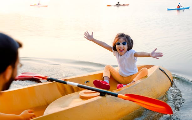 Toddler kayaking on a calm pond with arms outstretched, wearing sunglasses.