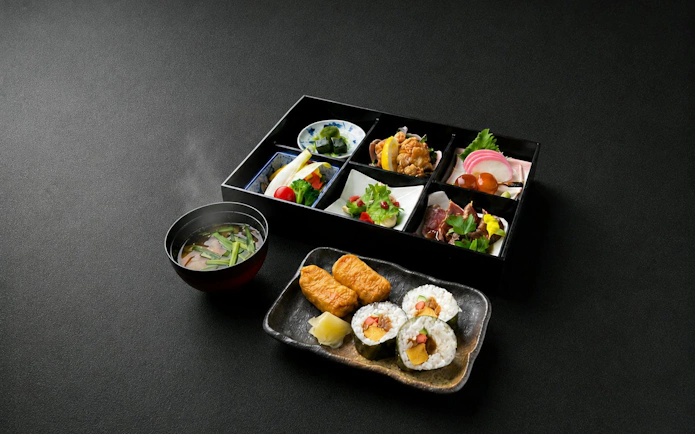 Japanese bento box with sushi rolls and miso soup on a black surface.