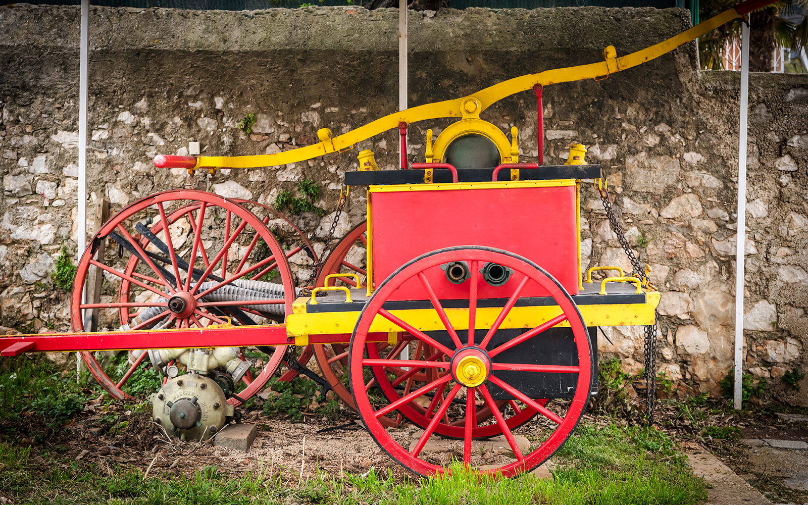Old vintage manual fire pump with red and yellow wheels against a stone wall.