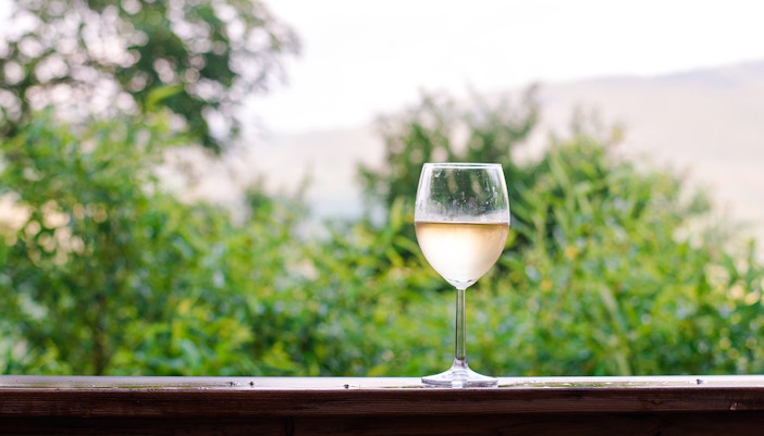 Glass of white wine on a wooden railing with a lush green background.