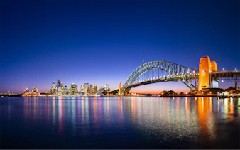 Sydney Harbour Bridge and Opera House illuminated at night with city skyline reflected in the water.