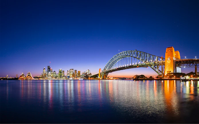 Sydney Harbour Bridge and Opera House illuminated at night with city skyline reflected in the water.