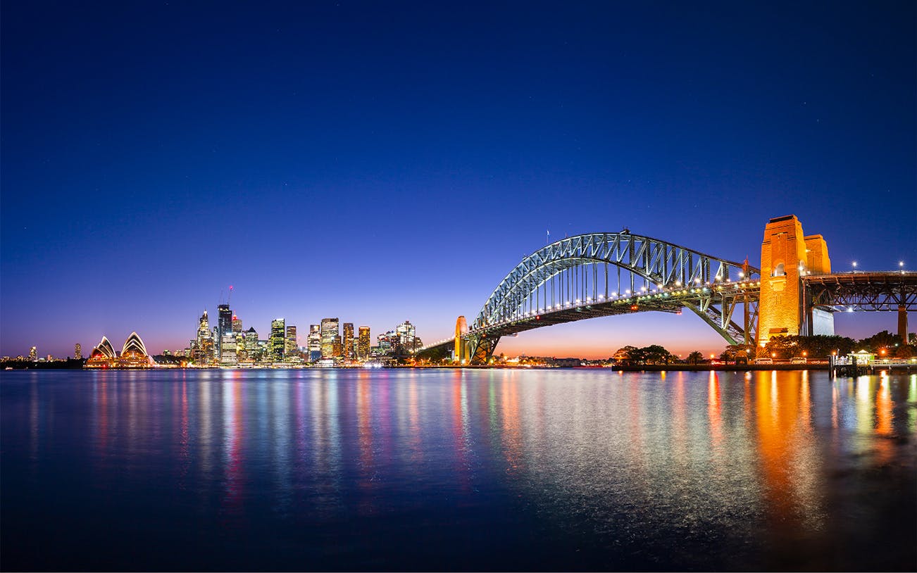Sydney Harbour Bridge and Opera House illuminated at night with city skyline reflected in the water.