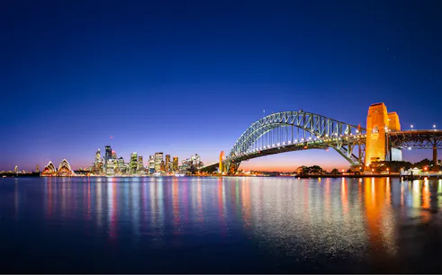 Sydney Harbour Bridge and Opera House illuminated at night with city skyline reflected in the water.