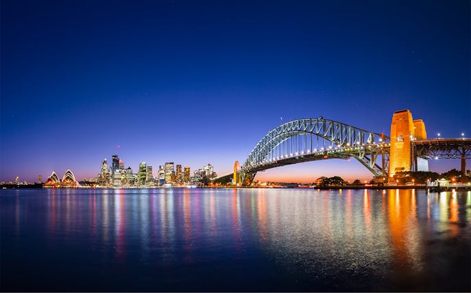 Sydney Harbour Bridge and Opera House illuminated at night with city skyline reflected in the water.