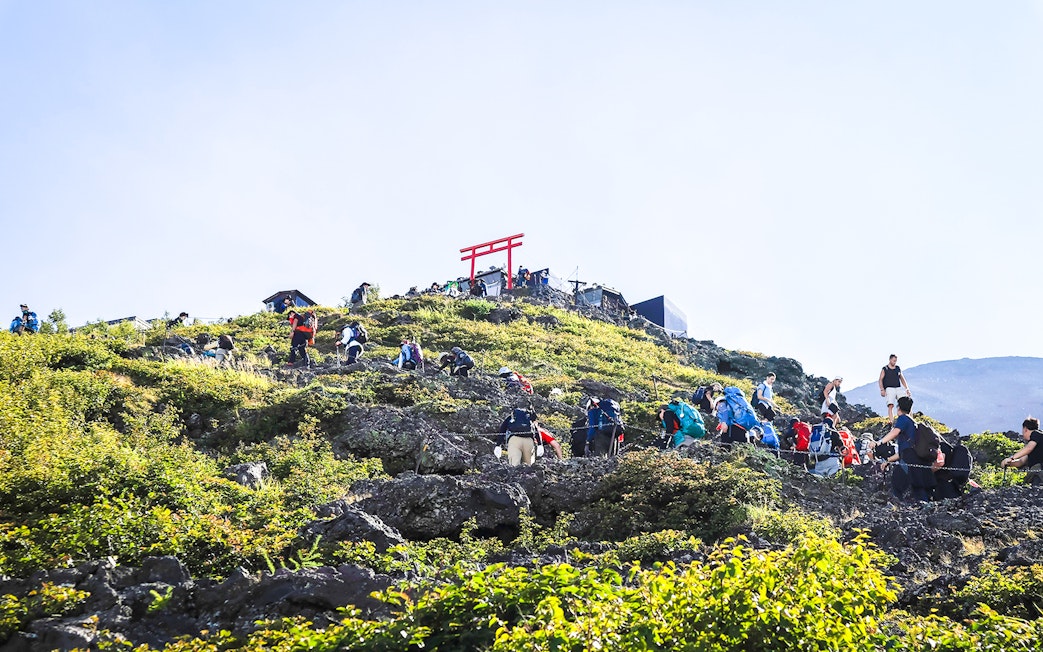 Hikers ascending Mount Fuji trail with distant red Torii gate.