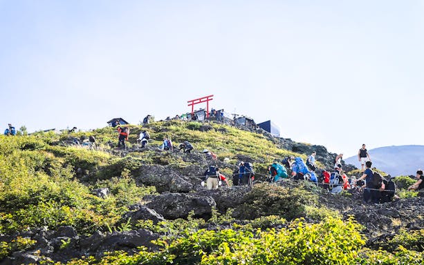 Hikers ascending Mount Fuji trail with distant red Torii gate.