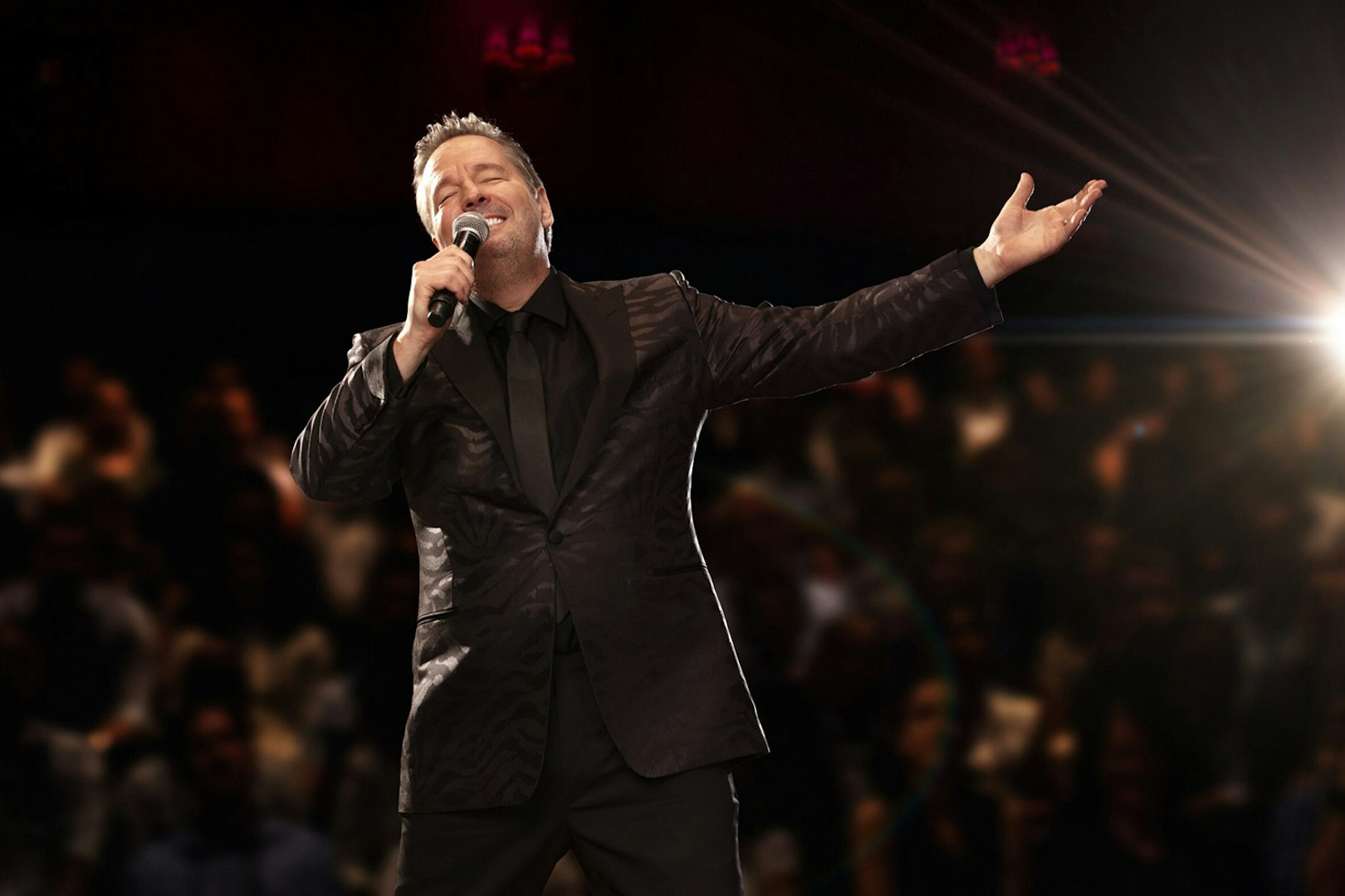 Entertainer performing on stage with microphone, spotlight, and audience in background.