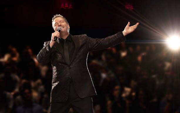 Entertainer performing on stage with microphone, spotlight, and audience in background.