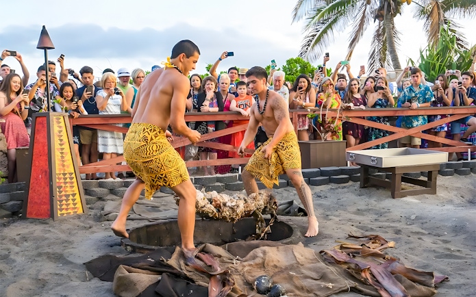 Men in traditional attire preparing a pig for roasting at Germaine's Luau, Hawaii, with a crowd watching.