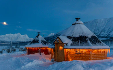 Wooden huts with lights in snowy Tromso landscape, reindeer grazing nearby.