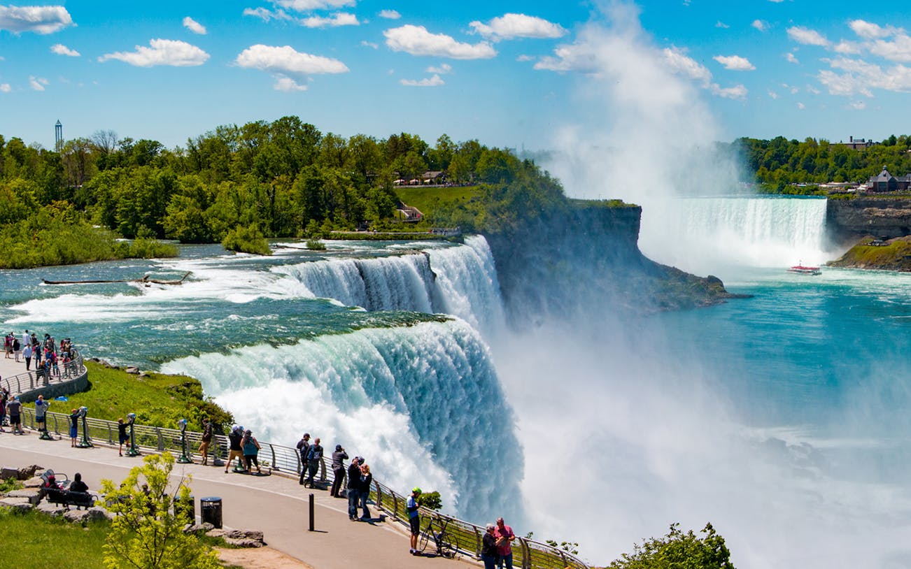 Tourists viewing Niagara Falls from observation deck with mist and boat below.