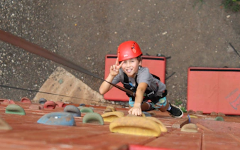 Child climbing wall on Adventure Course Extreme, wearing safety gear, showing peace sign.