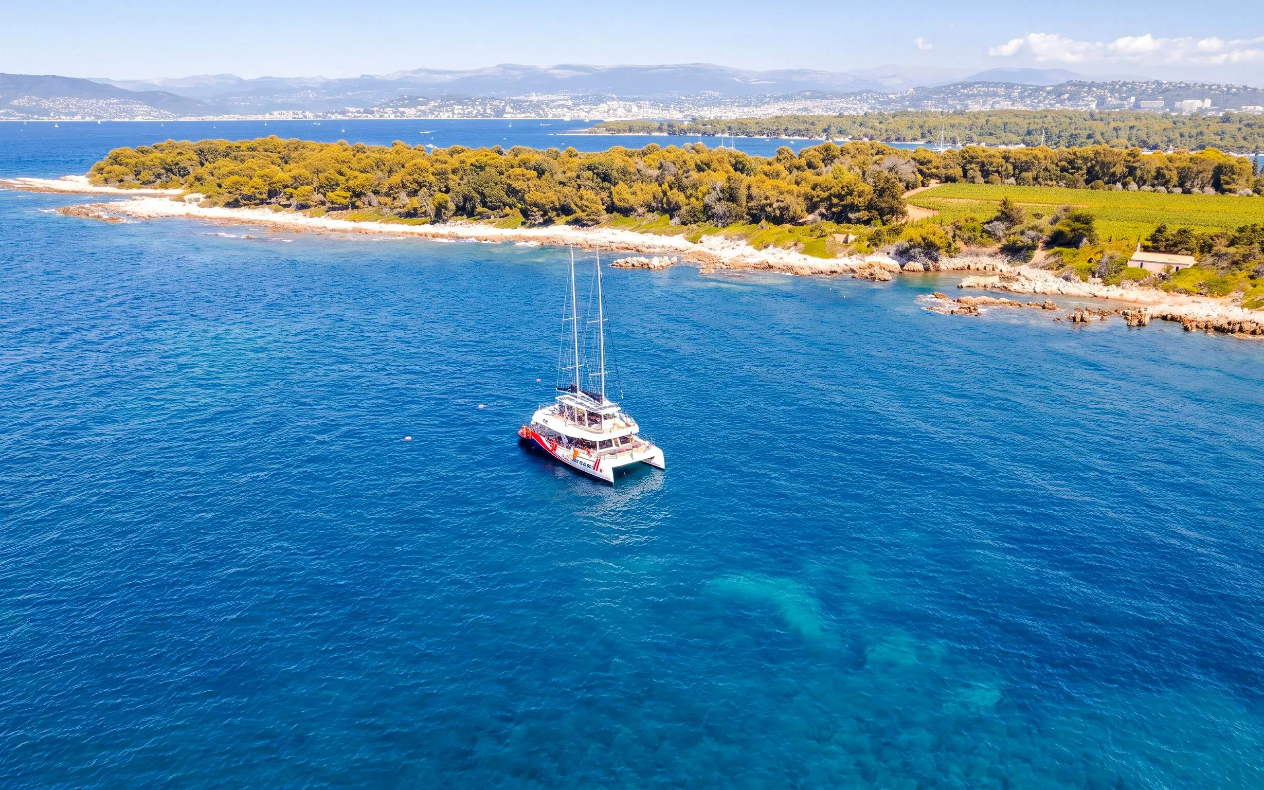 Catamaran cruising near Lérins Island, Marina Bay of Angels, with coastal landscape.