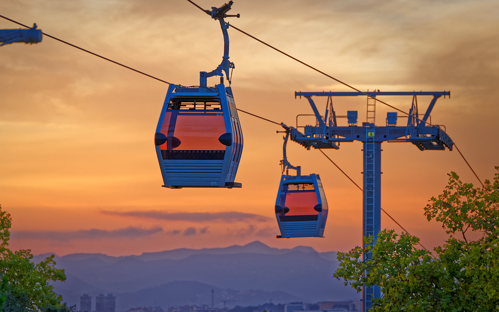 Cable cars at sunset over Barcelona, Spain, with cityscape and mountains in the background.