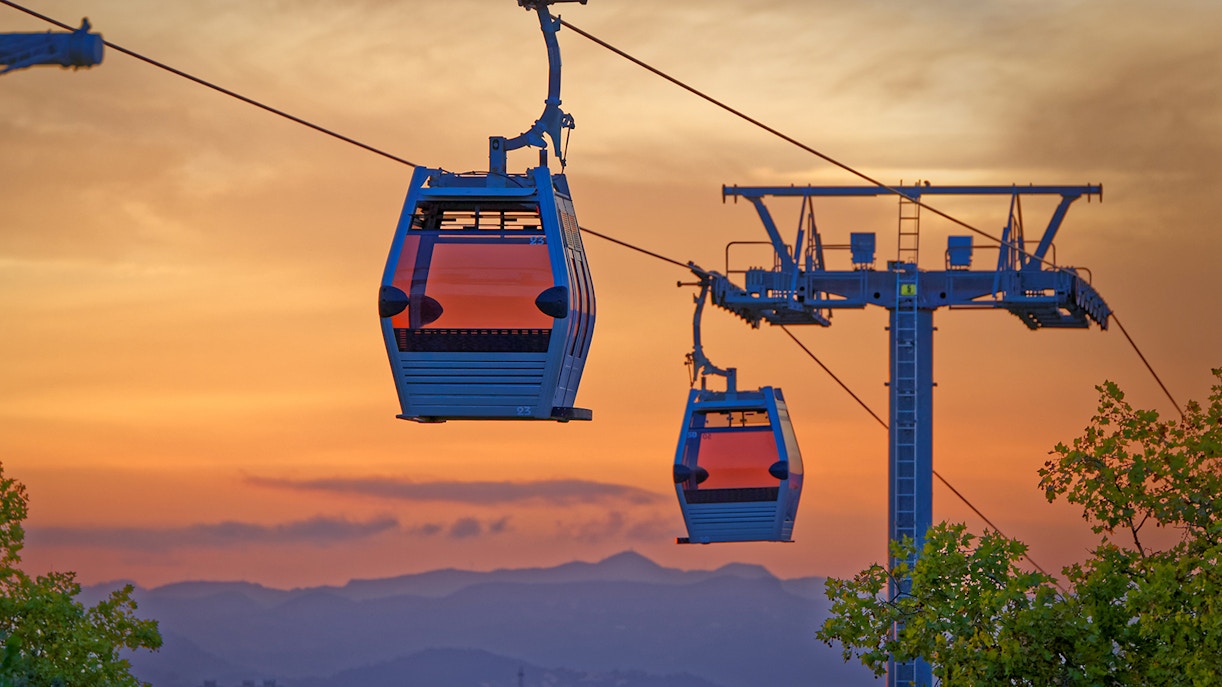 Cable cars at sunset over Barcelona, Spain, with cityscape and mountains in the background.