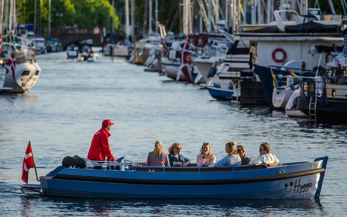 Guests on a guided boat tour in Copenhagen canal with Danish flag and moored boats.