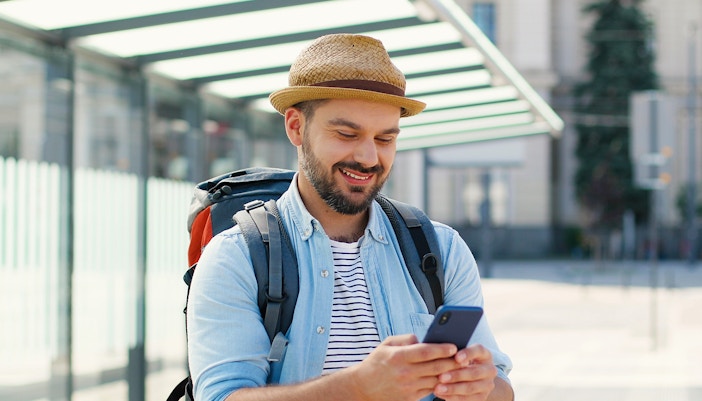 Man with a backpack using phone while waiting for a bus in a city setting.
