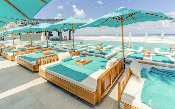 Daybeds at Atlas Beach Club, Bali overlooking the ocean with palm trees and clear skies.