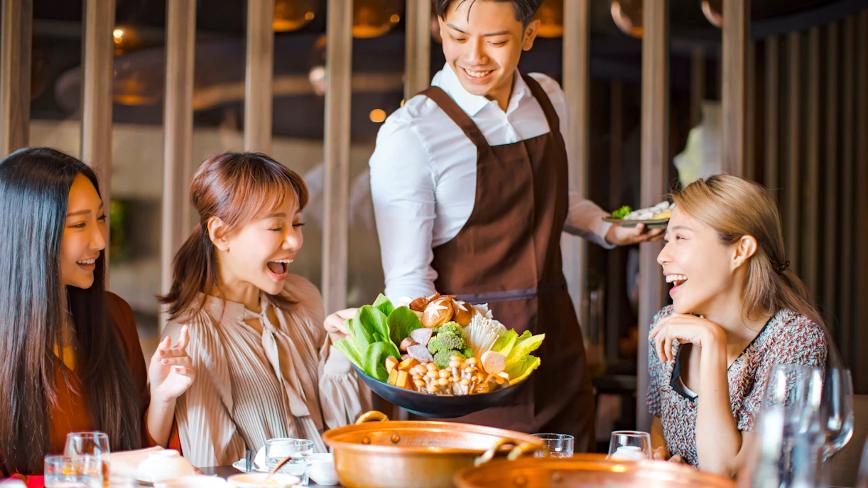 Waiter serving vegetables for hot pot to a group of friends in a restaurant.